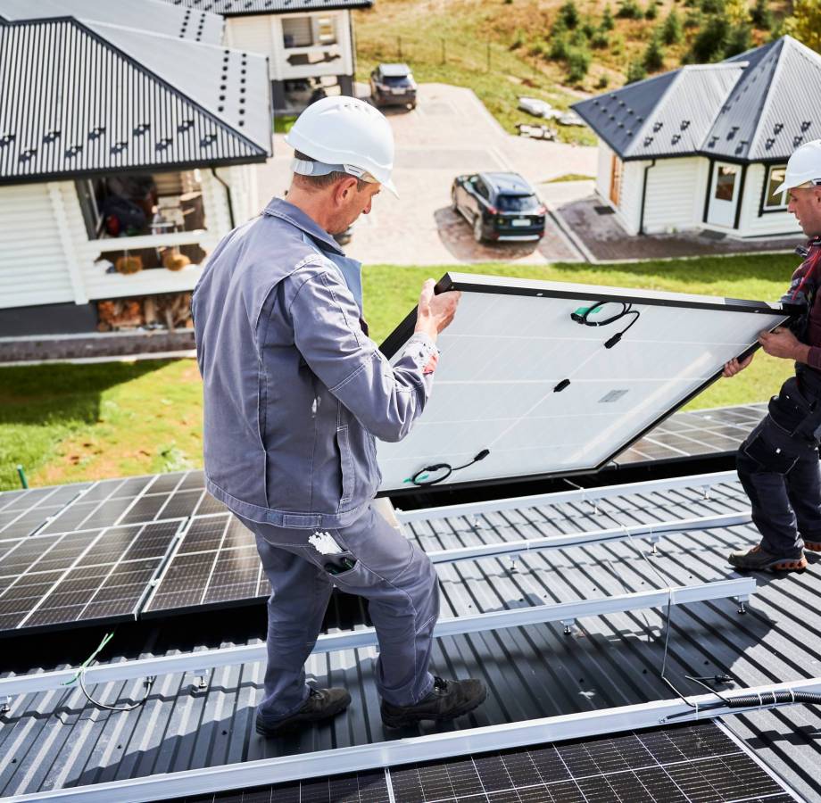 Mounters building solar panel system on roof of house. Men workers in helmets carrying photovoltaic solar module outdoors. Concept of alternative and renewable energy.